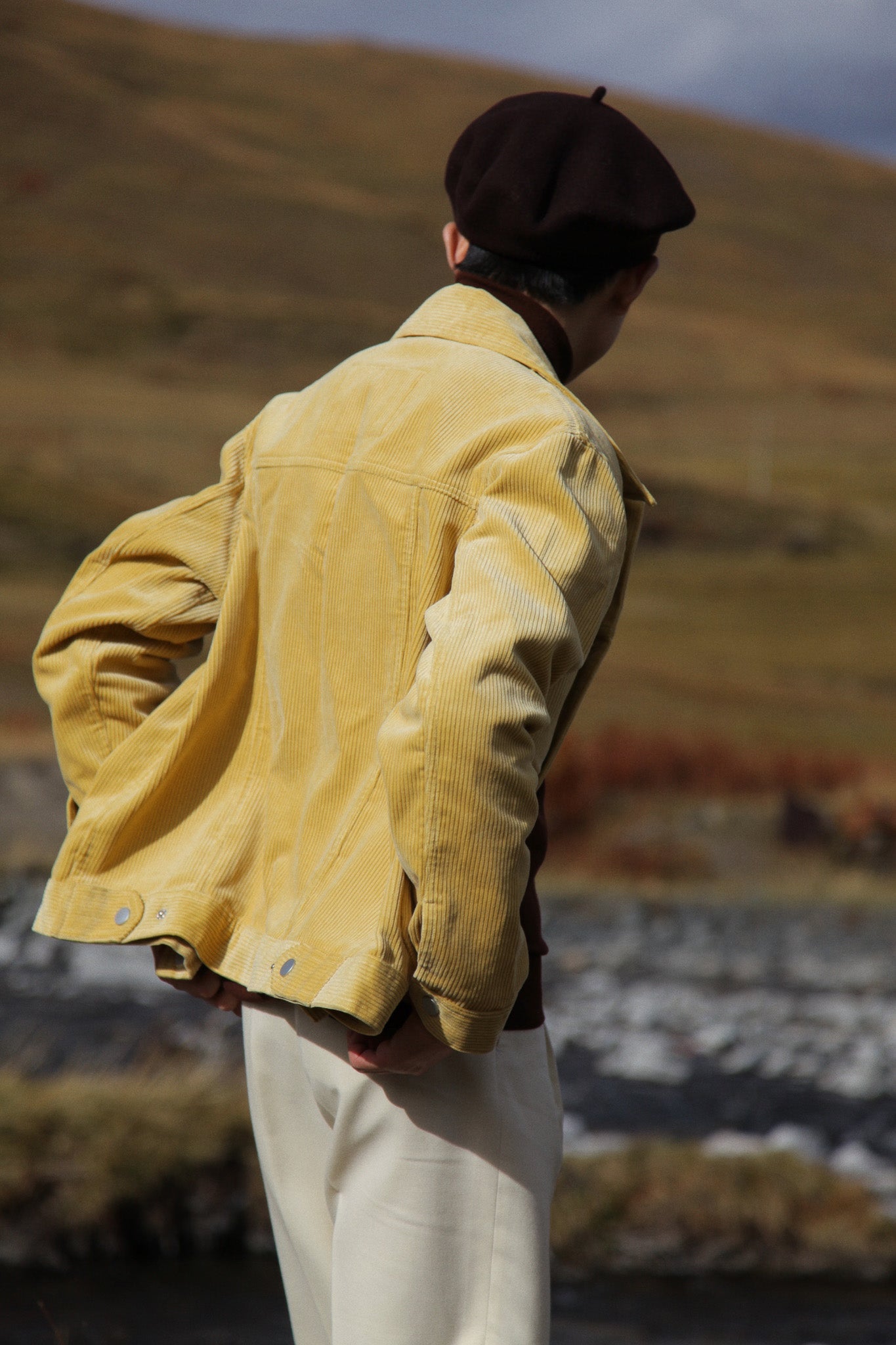 Person wearing a yellow jacket and dark hat standing in a field with mountains in the background #color_creamy-yellow