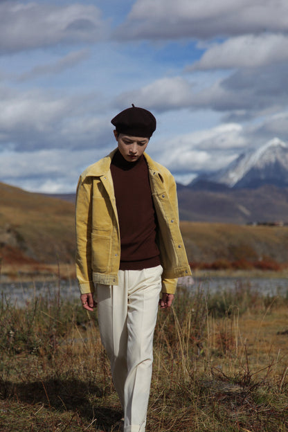 Person wearing a yellow jacket and white pants standing in a field with mountains in the background. Moonland men in corduroy workwear front view #color_creamy-yellow