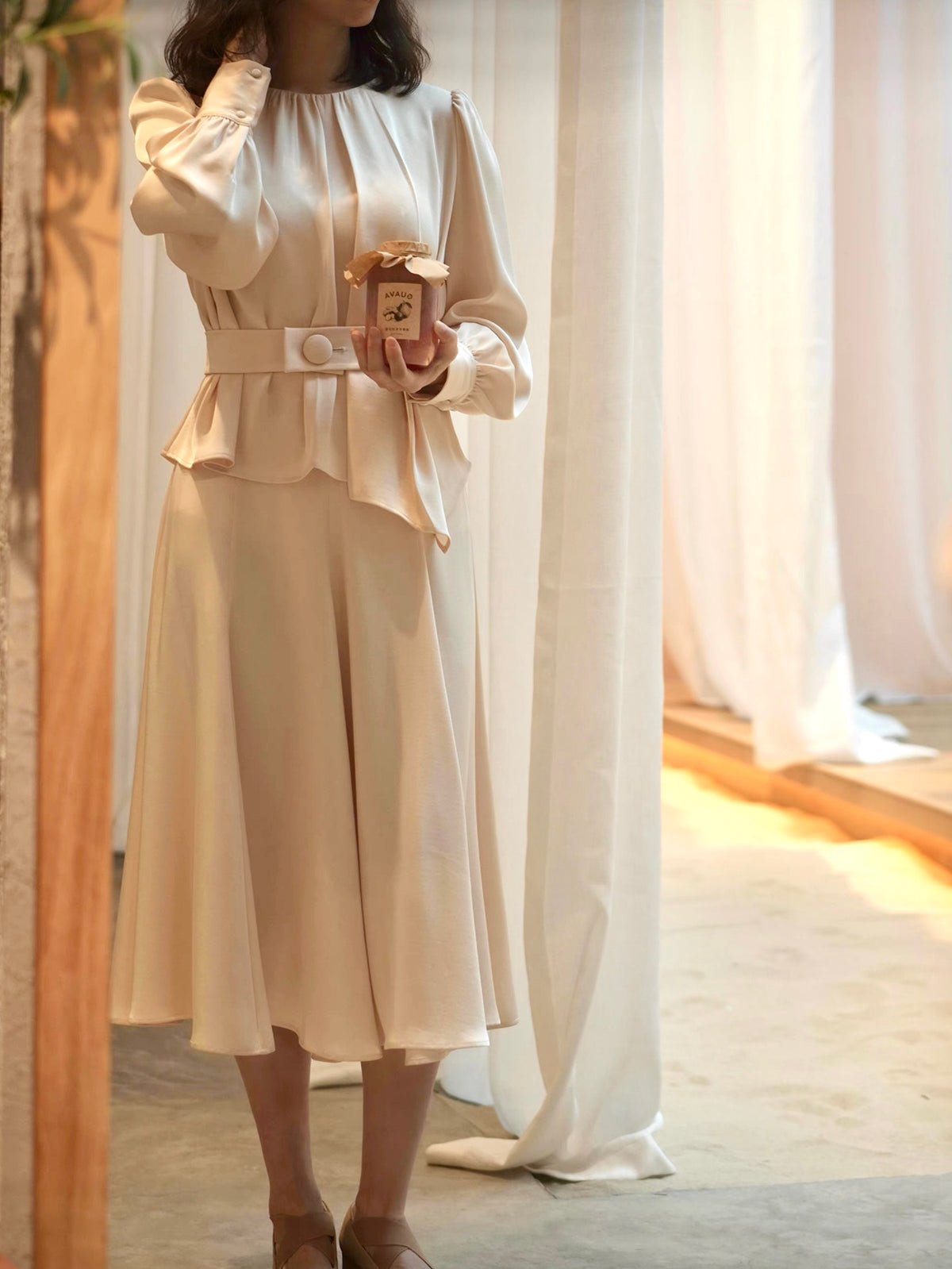 Moonland Silk Ripple Pleat Blouse, in champagne color, front view, gathered round neckline, long puff sleeves with volume, cuff with covered round buttons, pleated peplum with self belt at the front and a large covered button, paired with midi skirt to calf length, A-line silhouette with soft drape