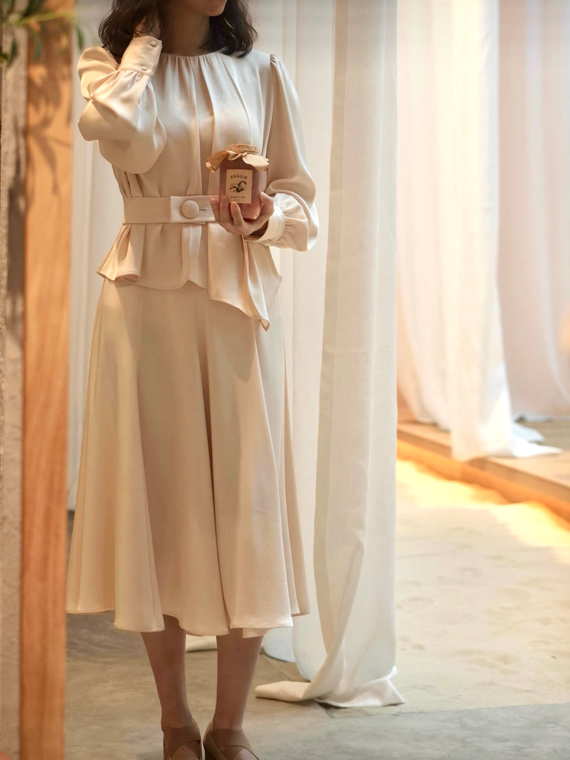 Moonland Silk Ripple Pleat Blouse, in champagne color, front view, gathered round neckline, long puff sleeves with volume, cuff with covered round buttons, pleated peplum with self belt at the front and a large covered button, paired with midi skirt to calf length, A-line silhouette with soft drape