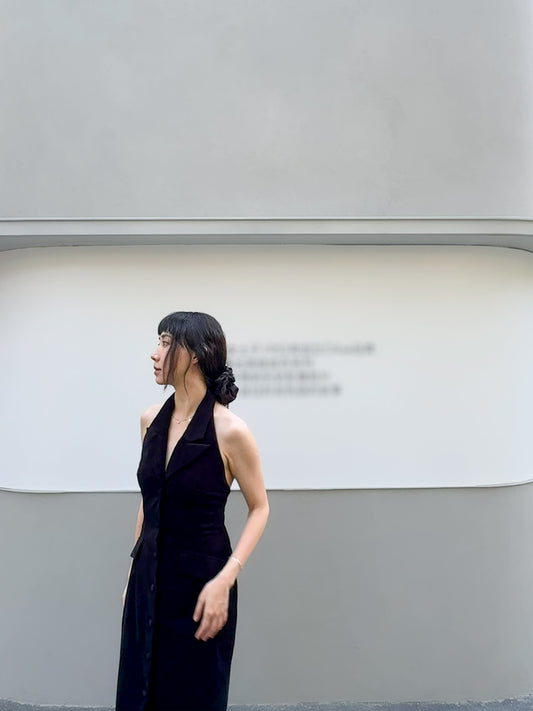 Moonland Smoking velvet halter dress, side front view by a wall, suit front with tailored lapel and jetted flap pockets, in black silk velvet, sheath silhouette