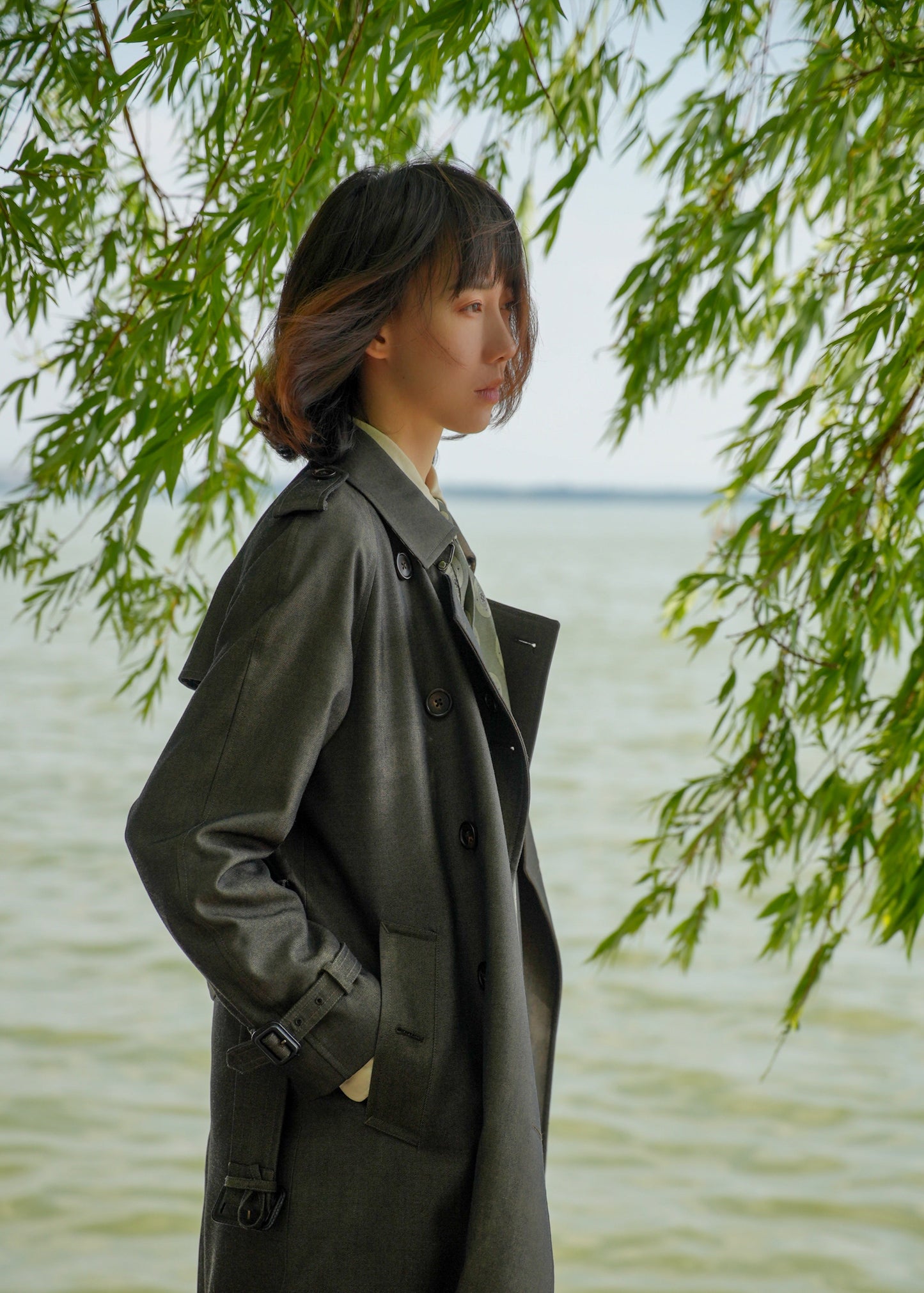 Moonland dark green trench coat with detachable down layer, double breasted with epaulettes and sleeve straps, side view under willow. #color_cypress-green