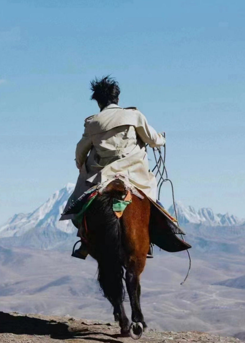 Person wearing a treach coat riding a horse with mountains in the background  #color_khaki