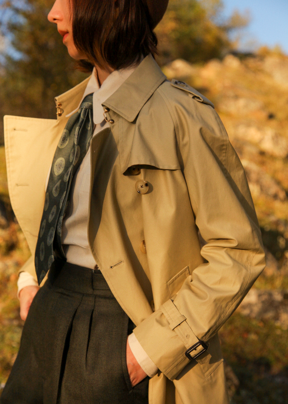 Person wearing a khaki trench coat in an outdoor setting with blurred natural background #color_khaki