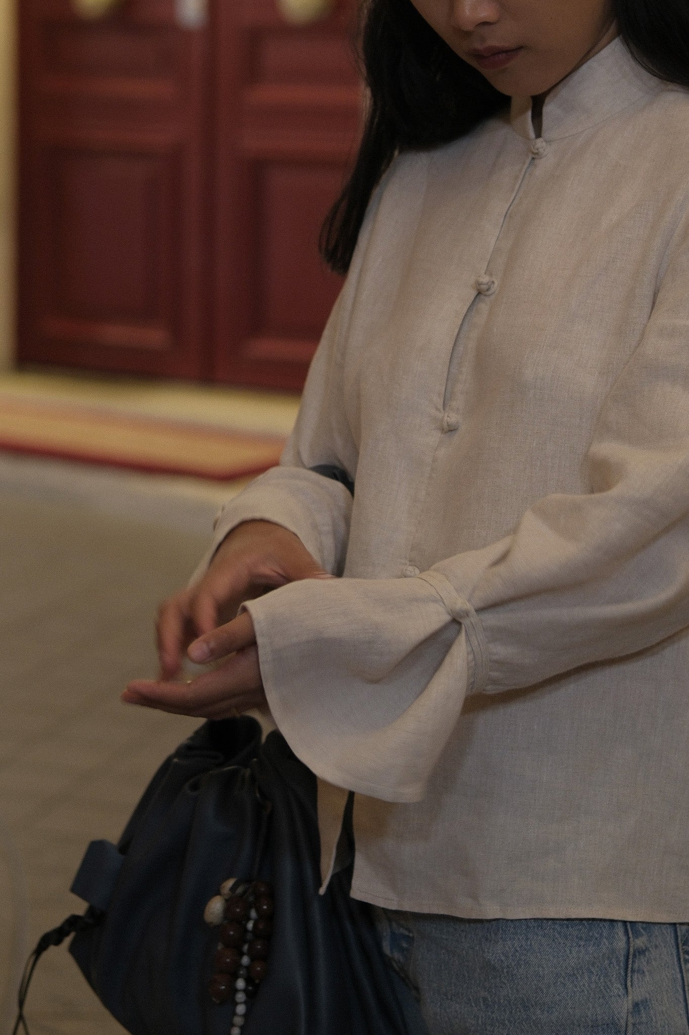 A woman wearing a light beige linen long-sleeve blouse with a mandarin collar and buttoned front, paired with blue jeans and holding a dark navy drawstring bag. #color_off-white