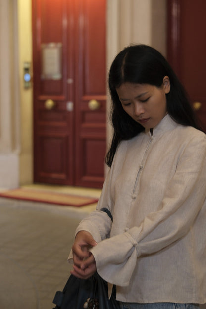 A woman wearing a light beige long-sleeve blouse with a high collar and front buttons, standing indoors near a dark red wooden door and holding a dark bag. #color_off-white