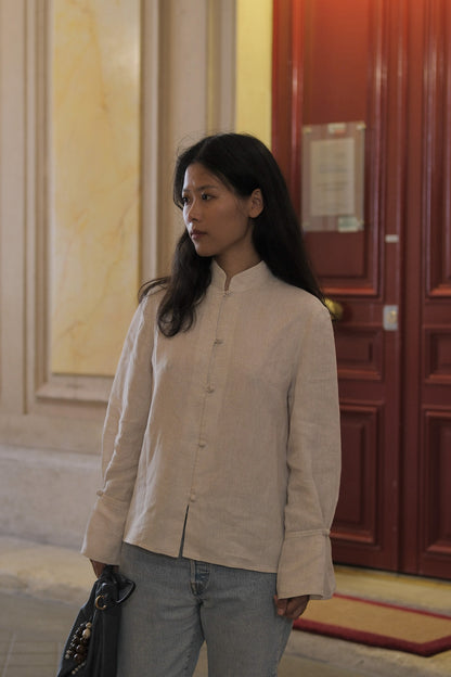 A woman wearing a light beige linen button-up jacket with a mandarin collar and light gray jeans, standing in an indoor hallway with a red wooden door in the background. #color_off-white
