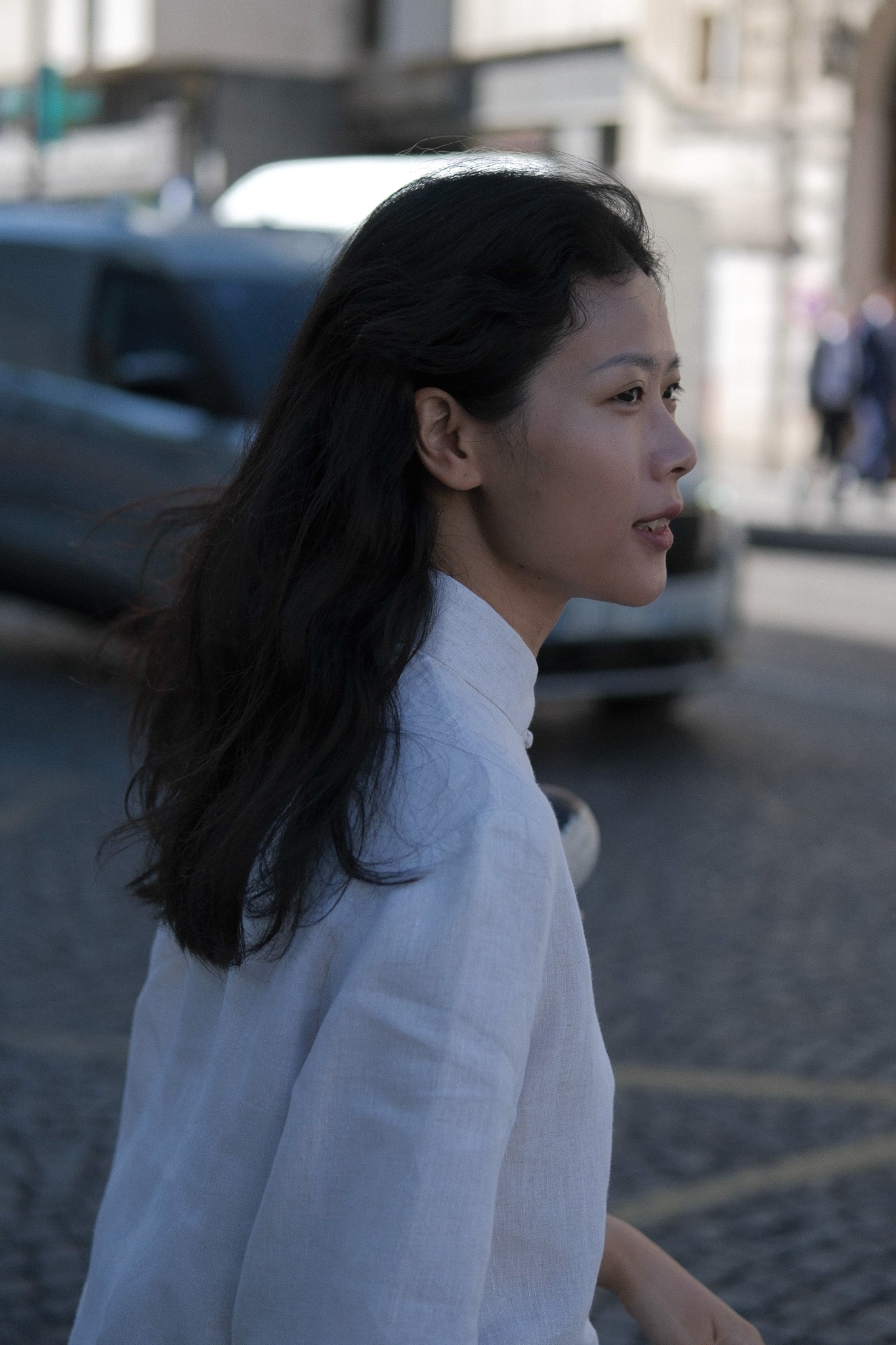 A woman in a white high-neck linen shirt walking outdoors on a cobblestone street, captured in a side profile with a blurred city background. #color_off-white