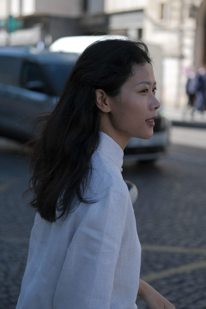 A woman in a white high-neck linen shirt walking outdoors on a cobblestone street, captured in a side profile with a blurred city background. #color_off-white